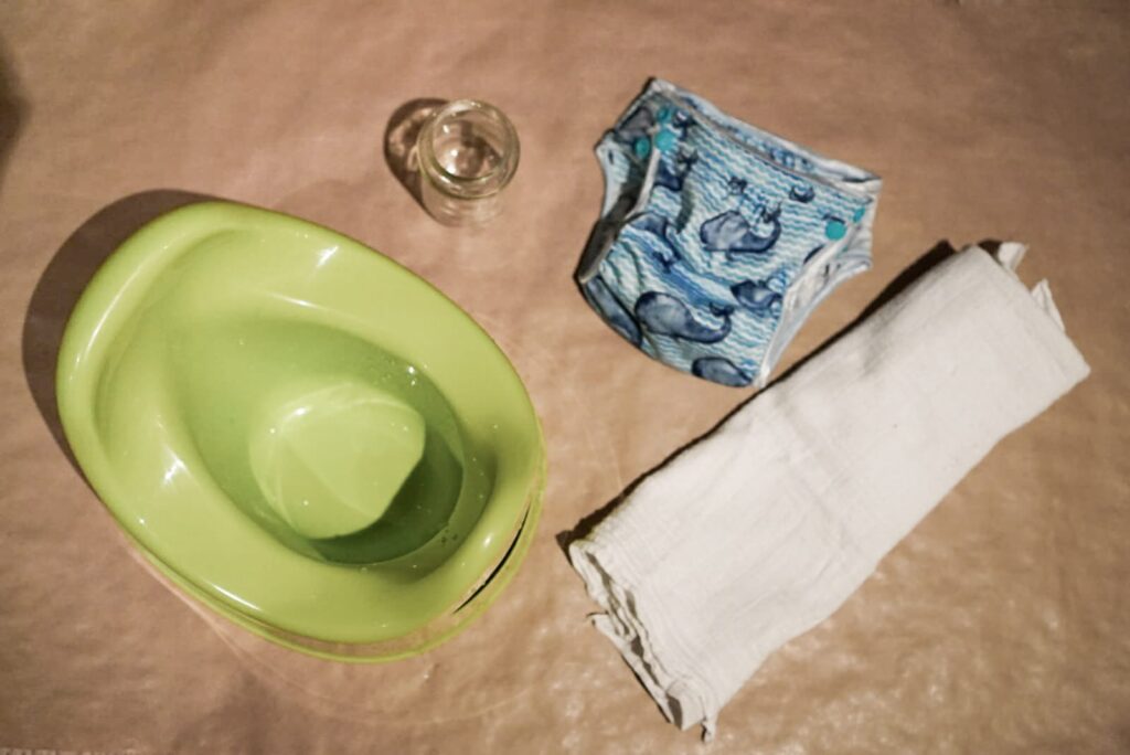 Auf dem Bild sind ein Töpfchen, ein Schraubglas, eine Abhaltewindel und Mullwindel zu sehen. Alles was man zum Abhalten braucht